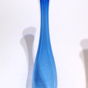 Op de afbeelding: Een hoge, slanke glazen vaas in een levendige kobaltblauwe kleur. De vaas heeft een smalle, langwerpige hals die geleidelijk breder wordt naar een afgeronde, bolvormige basis. Het glas is glad en doorschijnend en vangt het licht op.
