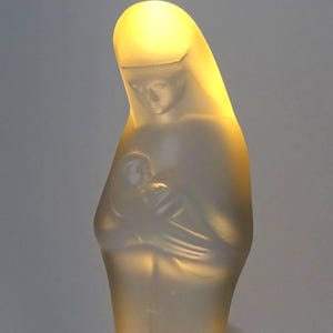 May include: A frosted glass sculpture of a woman holding a child. The sculpture is illuminated from within, creating a soft, warm glow. The woman is draped in a veil, and the child is cradled in her arms. The sculpture has a smooth, matte finish.