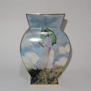 Op de afbeelding: Een decoratieve vaas met een schilderij van een vrouw met een groene paraplu in een veld. De vaas heeft een gouden rand en een unieke vorm. Het kunstwerk toont een vrouw in een witte jurk, tegen een blauw-witte lucht.