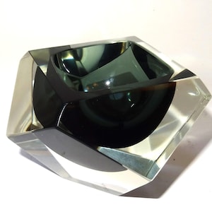 Puede incluir: Un cenicero geométrico de vidrio transparente con interior gris oscuro. El cenicero tiene un diseño facetado con una abertura cuadrada y un cuenco profundo. El vidrio es grueso y parece soplado a mano.