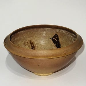 Może przedstawiać: Ręcznie wykonana ceramiczna miska z szerokim brzegiem i zaokrąglonym korpusem. Misa ma ciepłą, ziemistą paletę kolorów z odcieniami brązu i beżu. Wnętrze ma teksturowaną powierzchnię z ciemnobrązowymi akcentami.