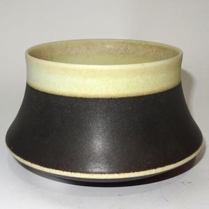 Może przedstawiać: Ceramiczna miska o szerokim, rozkloszowanym kształcie. Miska ma ciemnobrązową dolną część i jasnobeżową górną część. Obręcz jest w jaśniejszym beżowym kolorze. Miska jest prawdopodobnie używana do serwowania lub ekspozycji.