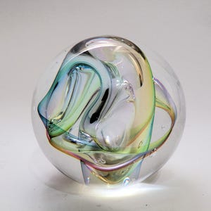 Op de afbeelding: Een heldere glazen presse-papier bol met een intern wervelend ontwerp van iriserende linten in tinten blauw, groen en geel. De massieve glazen bol bevat kleine luchtbellen die in het heldere materiaal zweven, wat zorgt voor een reflecterend oppervlak.
