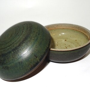 Puede incluir: Un cuenco de cerámica hecho a mano con tapa. El cuenco es de color verde oscuro con un interior moteado y una base verde más clara. La tapa ovalada tiene un diseño en espiral. El borde es de color marrón claro.