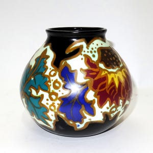 May include: A small, round ceramic vase with a black rim and base. The vase is decorated with colorful leaf patterns in teal, blue, red, and yellow, outlined in gold against a white and black background. The vase has a glossy finish.