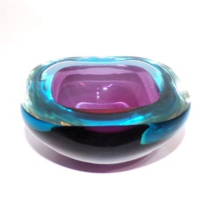 Könnte beinhalten: Eine dickwandige Glasschale mit einem tiefvioletten Innenkern, der von einer leuchtend türkisfarbenen Schicht umhüllt ist, und einem äußeren Rand aus klarem Glas. Die Schale hat eine abgerundete, organische Form mit einer glatten, polierten Oberfläche.