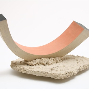 Puede incluir: Una escultura de cerámica con una forma curva de color naranja que descansa sobre una base blanca rugosa.