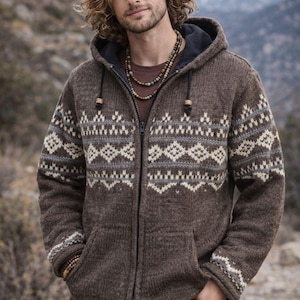 Könnte beinhalten: Braune Kapuzenjacke mit Reißverschluss und geometrischem Muster in Weiß und Grau. Die Jacke hat eine Kapuze mit Kordelzug und zwei Vordertaschen. Die Person im Bild trägt ein braunes Hemd und blaue Jeans. Im Hintergrund ist ein Gebirge zu sehen.