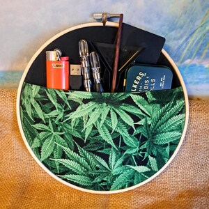 May include: A round, black fabric organizer with a pocket featuring a green cannabis leaf pattern. It holds an orange lighter, USB drive, metal tools, a small tin, and a wooden stick. The organizer is held in a wooden embroidery hoop.