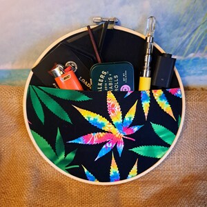 May include: A round, wooden embroidery hoop with a black fabric pocket featuring a tie-dye cannabis leaf design. The pocket holds a lighter, a small tin, and a vape pen. The background is a beach scene.