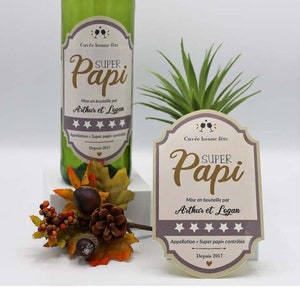 May include: A green wine bottle with a label that says "Super Papi" and is decorated with stars. The label also says "Mise en bouteille par Arthur et Legan" and "Appellation Super Papi contr&ocirc;l&eacute;e." The bottle is sitting on a table with a small plant and a decorative fall arrangement.