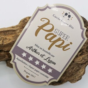 May include: A light brown wooden plaque with a white and purple label that reads "Cuv&eacute;e bonne f&ecirc;te SUPER Papi Mise en bouteille par Arthur et Logan Appellation Super papi contr&ocirc;l&eacute;e Depuis 2017".