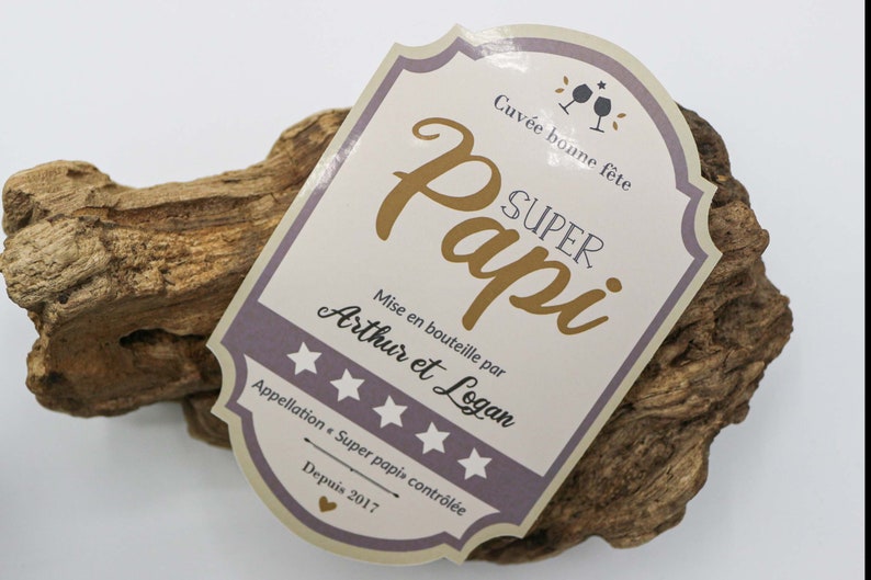 May include: A light brown wooden plaque with a white and purple label that reads "Cuv&eacute;e bonne f&ecirc;te SUPER Papi Mise en bouteille par Arthur et Logan Appellation Super papi contr&ocirc;l&eacute;e Depuis 2017".