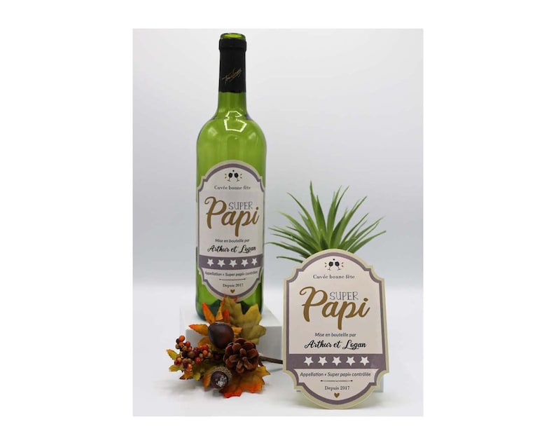 May include: A green wine bottle with a label that says "Super Papi" and is decorated with stars. The label also says "Mise en bouteille par Arthur et Legan" and "Appellation Super Papi contr&ocirc;l&eacute;e." The bottle is sitting on a table with a small plant and a decorative fall arrangement.