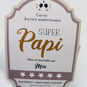 May include: A light brown and purple card with a wine bottle label design. The text reads "Cuv&eacute;e Joyeux anniversaire SUPER Papi Mise en bouteille par Mia Appellation "Super papi" contr&ocirc;l&eacute;e Depuis 2021".