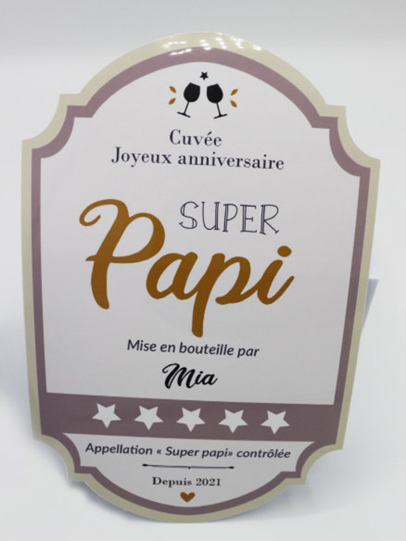 May include: A light brown and purple card with a wine bottle label design. The text reads "Cuv&eacute;e Joyeux anniversaire SUPER Papi Mise en bouteille par Mia Appellation "Super papi" contr&ocirc;l&eacute;e Depuis 2021".