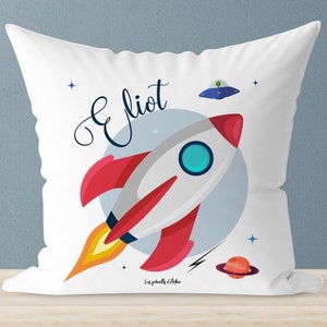 Può includere: Cuscino bianco con un razzo rosso e bianco, un UFO blu e verde e il nome "Elliot" in corsivo. Il cuscino ha uno sfondo bianco con stelle e un pianeta rosso.