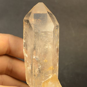 Puede incluir: Un cristal de cuarzo transparente con una punta puntiaguda y una base plana. El cristal mide aproximadamente 12,5 cm de alto y tiene una textura natural y rugosa.