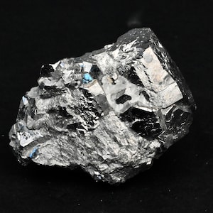 Può includere: Un gruppo di cristalli di colore grigio metallico con una superficie riflettente. I cristalli hanno una forma geometrica affilata e sono disposti in uno schema casuale.