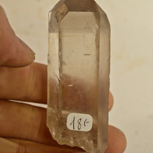 Può includere: Un cristallo di quarzo trasparente con una sfumatura marrone fumé. Il cristallo è appuntito in cima e ha una texture ruvida e naturale. Il cristallo è alto circa 12,5 cm ed è tenuto in una mano. Il cristallo ha un'etichetta con il numero 18C.