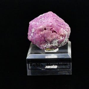 May include: A raw, vibrant pink ruby mineral specimen with crystalline textures and white inclusions, mounted on a clear, rectangular acrylic display base. The base features a black label with the printed text Rubis Macedoine.