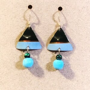 May include: A pair of triangle-shaped earrings with a black, gold, and light blue design. Each earring has a turquoise-coloured bead hanging from it.