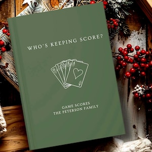 May include: A sage green hardcover book titled WHO'S KEEPING SCORE? with a white line art illustration of a royal flush of hearts playing cards. Below the cards, the text reads GAME SCORES and THE PETERSON FAMILY in white lettering.