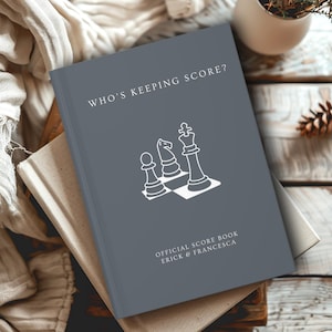 May include: A grey chess scorebook with the text "WHO'S KEEPING SCORE?" and a white graphic of chess pieces. The book is labelled "OFFICIAL SCORE BOOK ERICK & FRANCESCA". The book is resting on top of a beige book.