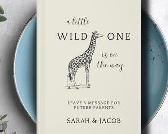 May include: A cream-colored guest book featuring a black line drawing of a giraffe in the center. The text reads: a little WILD ONE is on the way, LEAVE A MESSAGE FOR FUTURE PARENTS, and SARAH & JACOB. The book has a clean, minimalist design.