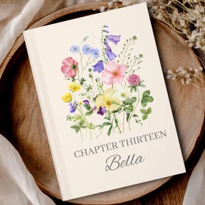 May include: A rectangular notebook with a cream-colored cover featuring a watercolor illustration of various wildflowers in shades of pink, yellow, blue, and purple. The text "CHAPTER THIRTEEN" and the name "Bella" are printed in gray below the flowers.