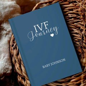 May include: A blue hardcover journal with white text that reads IVF Journey and Baby Johnson, featuring a small white heart icon between the words. The book has a smooth matte finish and is positioned at an angle on a textured surface.