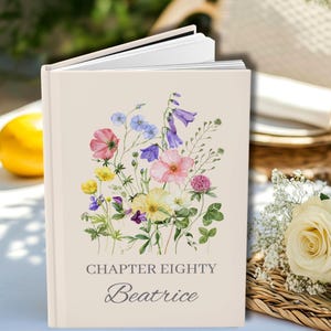 May include: A hardback book with a cream-coloured cover featuring a watercolour illustration of various wildflowers in shades of pink, yellow, blue, and purple. The text "CHAPTER EIGHTY" and the name "Beatrice" are printed in grey.