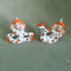May include: Two small porcelain clown figurines with orange and white polka dot costumes. The clowns are in different poses, one is sitting with legs crossed and the other is lying on its back with legs in the air.