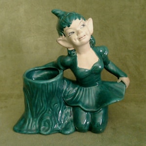 May include: A green ceramic figurine of a female elf with pointed ears, wearing a green dress and holding a green ceramic planter shaped like a tree stump.