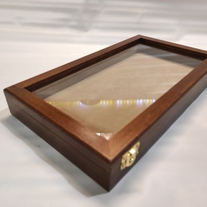Peut inclure: Une vitrine en bois brun foncé avec un dessus en verre et deux fermoirs en métal doré. La vitrine est vide et a une doublure en feutre brun clair.