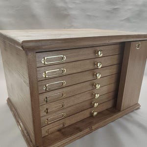 Könnte beinhalten: Ein Aufbewahrungsschrank aus Holz mit sieben horizontalen Schubladen, die mit goldenen Metall-Etikettenhaltern und kleinen runden goldenen Knöpfen versehen sind. Der Schrank hat ein vertikales Seitenteil mit einem goldenen Akzent.