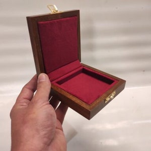 Può includere: Una piccola scatola di legno con un interno in velluto rosso e una chiusura in ottone. La scatola è aperta e l'interno è visibile.