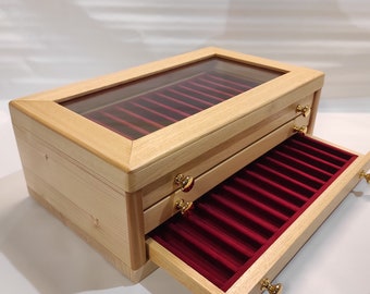 Könnte beinhalten: Eine Holzstiftebox mit Glasdeckel und zwei Schubladen. Die Schubladen sind mit rotem Filz ausgekleidet und haben goldfarbene Knöpfe. Die Box ist aus hellem Holz gefertigt und hat eine natürliche Oberfläche.