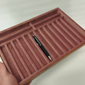 May include: A wooden tray with a burgundy velvet lining and multiple slots for storing pens or other small items. A black pen with the text "WATERMAN" is lying in one of the slots.