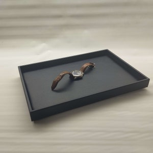 May include: A black rectangular tray with a brown leather watch band and a silver watch face.