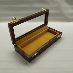 Op de afbeelding: Een donkerbruine houten doos met een glazen deksel en een gele fluwelen voering. De doos heeft twee messing sluitingen aan de voorkant en twee aan de zijkant.
