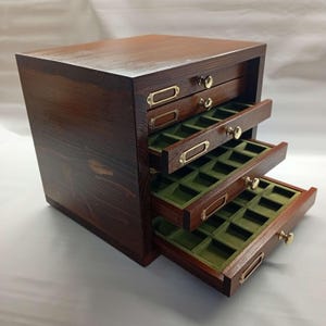 Puede incluir: Una caja de joyería de madera teñida oscura con tres cajones parcialmente abiertos. Los cajones tienen forro de fieltro verde y herrajes de latón. La caja es rectangular con una parte superior ligeramente inclinada.