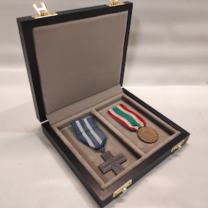 Puede incluir: Una caja de madera negra con un interior de terciopelo gris, que contiene dos medallas con cintas. Una medalla es una cruz plateada con una cinta azul y blanca, y la otra es una medalla redonda de bronce con una cinta verde, blanca y roja.