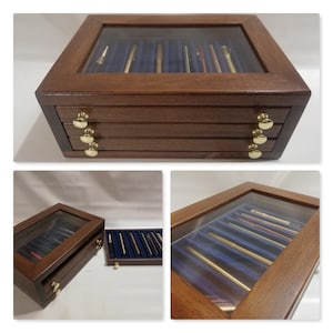 Könnte beinhalten: Eine Holzstiftebox mit Glasdeckel und drei Schubladen. Die Box ist mit Stiften gefüllt und hat goldfarbene Knöpfe.