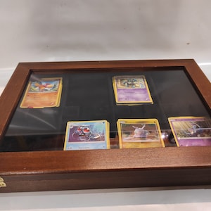 May include: A wooden display case with a glass top, showcasing a collection of Pokemon trading cards. The cards feature various Pokemon characters, including Charizard, Snorlax, and Venusaur.