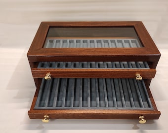 Puede incluir: Una caja de madera para bolígrafos con tapa de vidrio y dos cajones. Los cajones están abiertos y forrados con terciopelo azul. La caja tiene asas de latón.