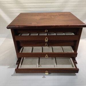 May include: A dark brown wooden jewelry box with four drawers and gold-colored handles. The top drawer is open, revealing a soft gray lining with compartments.