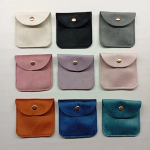 May include: A collection of nine square pouches arranged in a three-by-three grid. Each pouch features a soft, suede-like texture, a rounded flap, and a single gold-toned metal snap button closure. The colors include white, black, gray, dusty rose, lavender, light pink, burnt orange, royal blue, and teal.