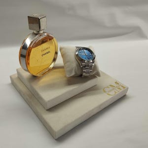 May include: A white display stand with the text "CM" and "Coins & More" in gold lettering. On the stand is a silver watch with a blue face and a clear glass bottle of perfume with a silver cap and the text "CHANCE" and "CHANEL" on the label.