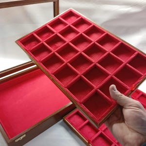May include: A wooden display case featuring a removable tray with multiple rectangular compartments. Both the interior of the main case and the tray are lined with bright red fabric. The tray is held at an angle, revealing its grid structure.
